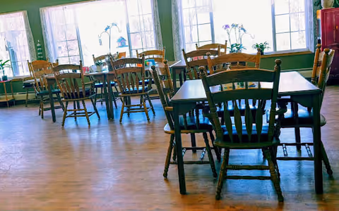 Dining room with several wooden tables and chairs arranged on a wood floor in front of large windows with potted plants.