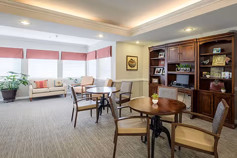 Well-lit communal room with round tables and chairs, a seating area by windows, and a wooden bookcase/entertainment cabinet.