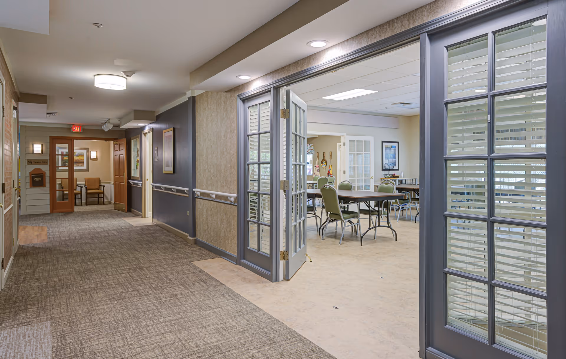 Interior view of a senior living facility hallway leading to a room with tables and chairs. The hallway has carpeted flooring, handrails on the walls, and framed artwork. The room to the right has glass-paneled double doors and contains several tables with green chairs arranged around them.