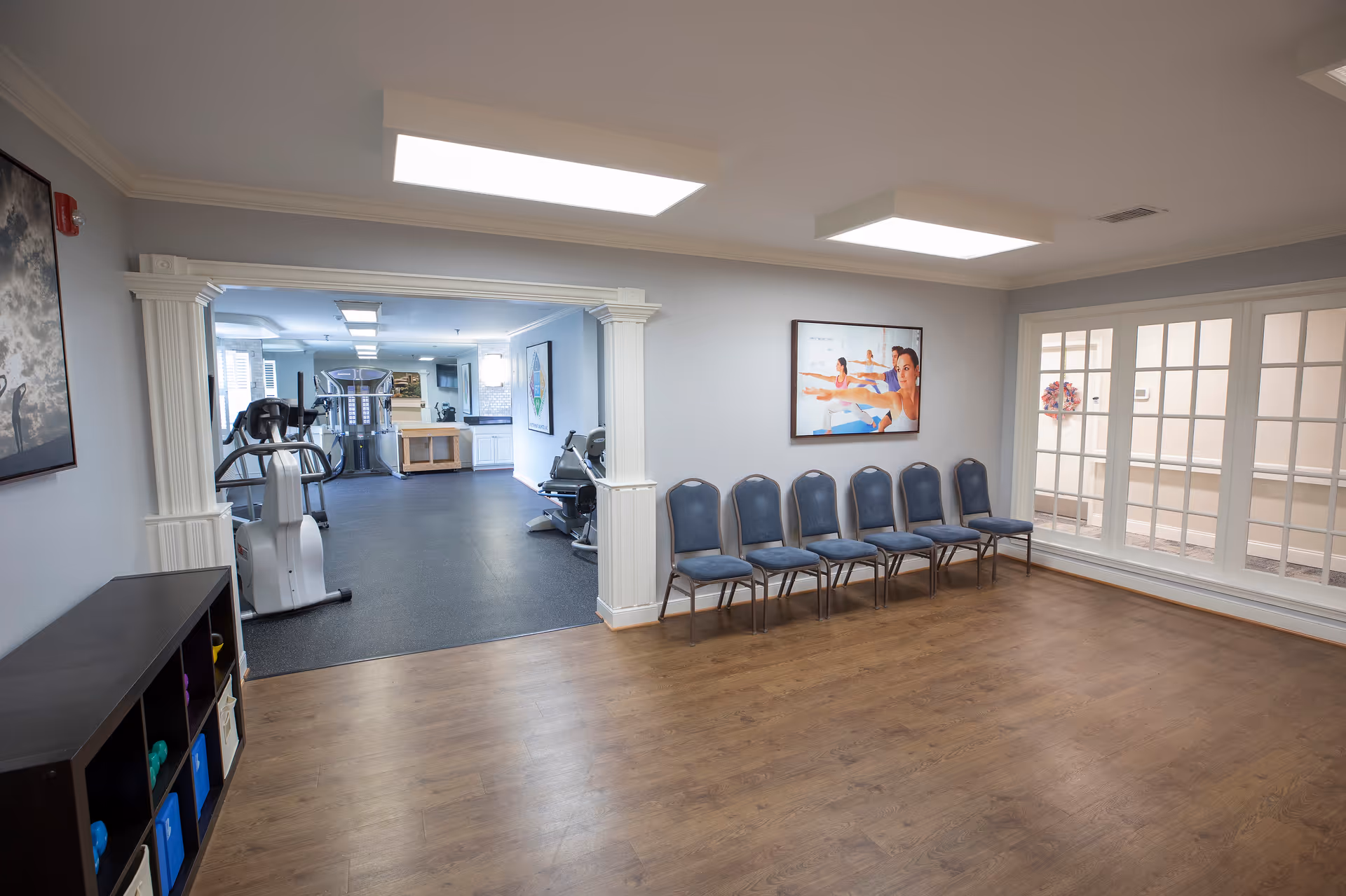 A spacious room with wooden flooring and a row of blue chairs lined up against a wall. The room opens into a fitness area equipped with exercise machines such as treadmills and stationary bikes. The walls are light-colored, and there is a large framed picture of people doing yoga on the wall above the chairs. Large windows with white frames are visible on the right side.