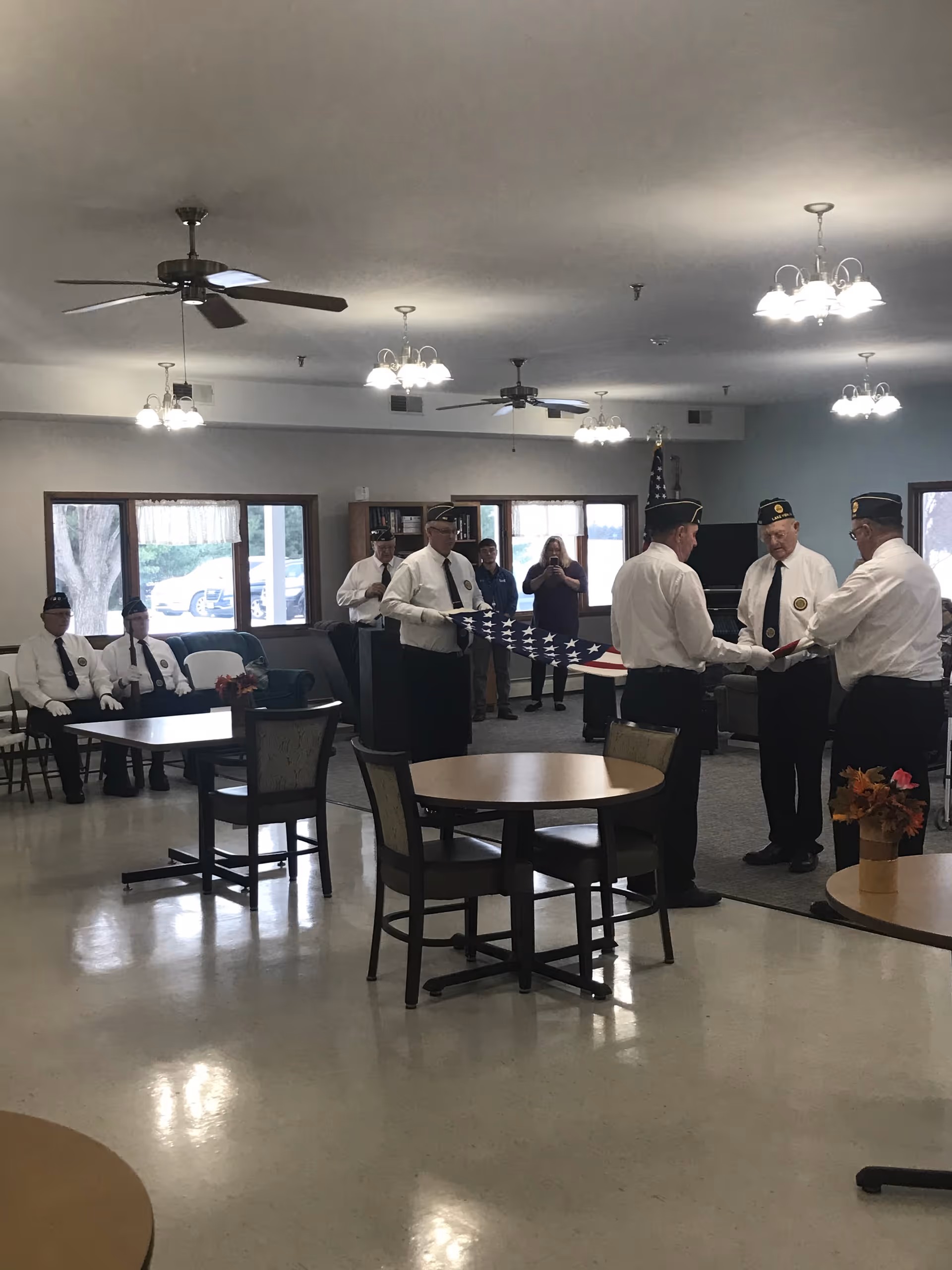 A group of men in uniform performing a flag folding ceremony inside a common room with tables, chairs, ceiling fans, and windows. Some people are seated while others stand and observe the ceremony.