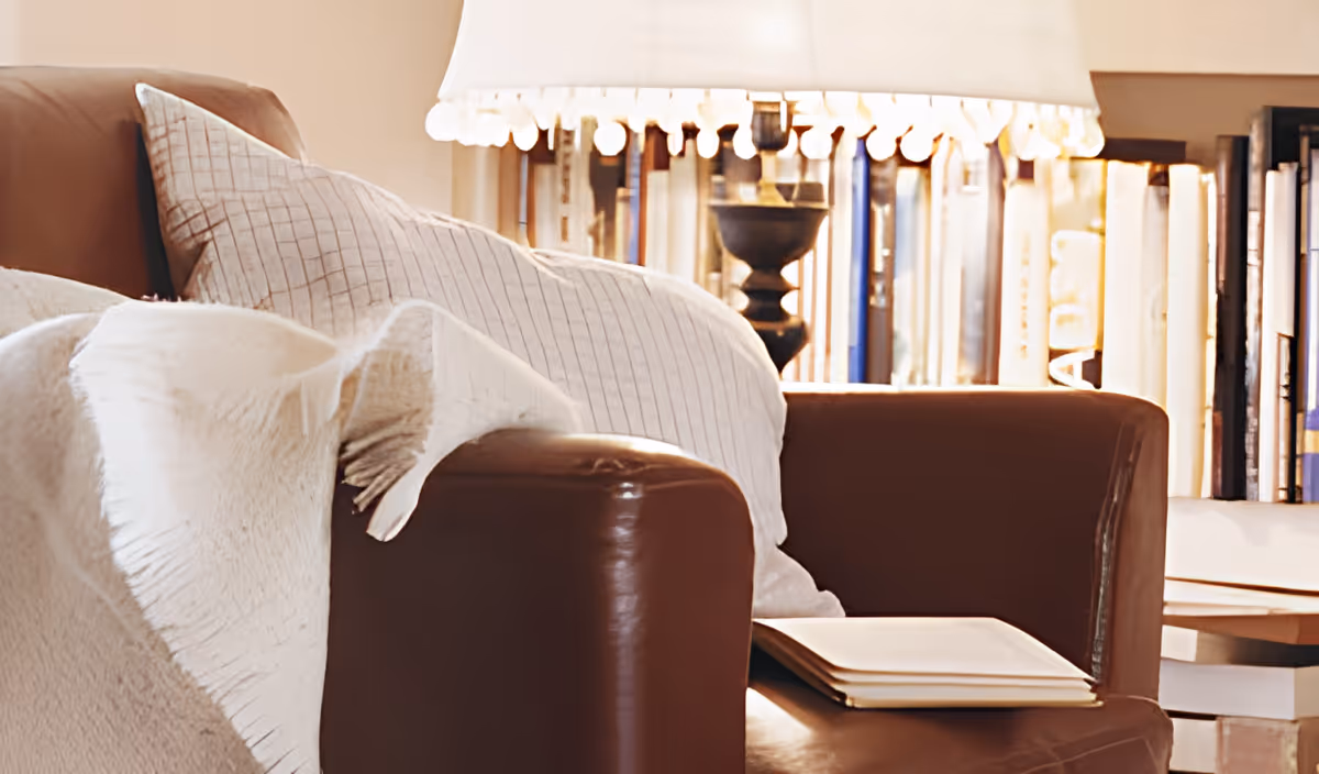 A cozy brown leather armchair with a white throw blanket and a white pillow with a grid pattern. Behind the chair is a bookshelf filled with books and a table lamp with a white lampshade.