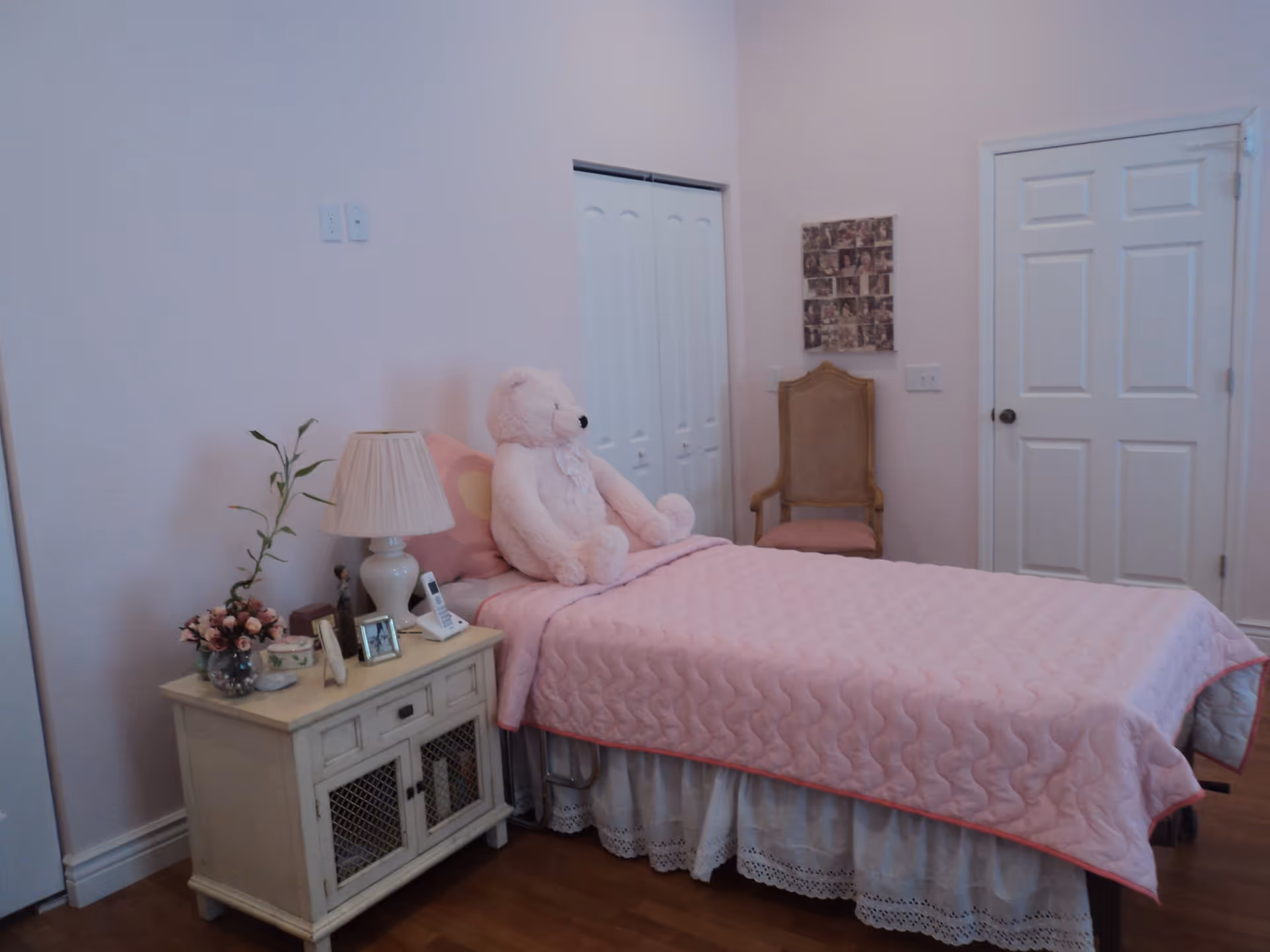 A bedroom with a single bed covered in a pink quilt and a large pink teddy bear sitting on the bed. Next to the bed is a white nightstand with a lamp, a small plant, framed photos, and other decorative items. There is a wooden chair with a pink cushion against the wall, a white closet door, and a white entry door in the background. The walls are painted light pink and the floor is wooden.