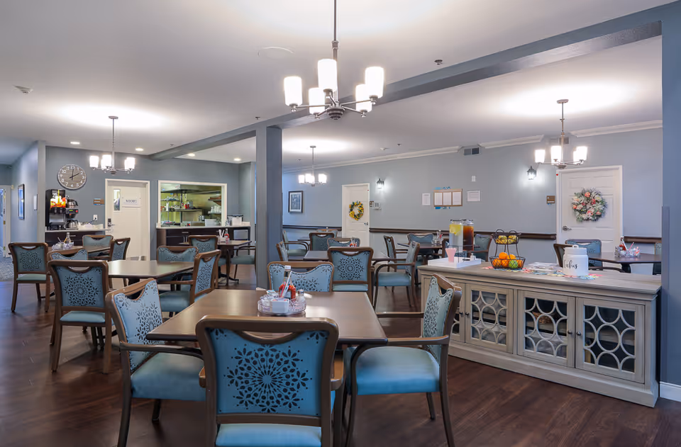 A spacious dining room in an assisted living facility with multiple wooden tables and blue cushioned chairs arranged neatly. The room has soft blue walls, dark wood flooring, and several ceiling light fixtures. A sideboard with refreshments, including pitchers of drinks and a fruit basket, is visible on the right. The kitchen area is partially visible in the background with a clock on the wall and a coffee station.