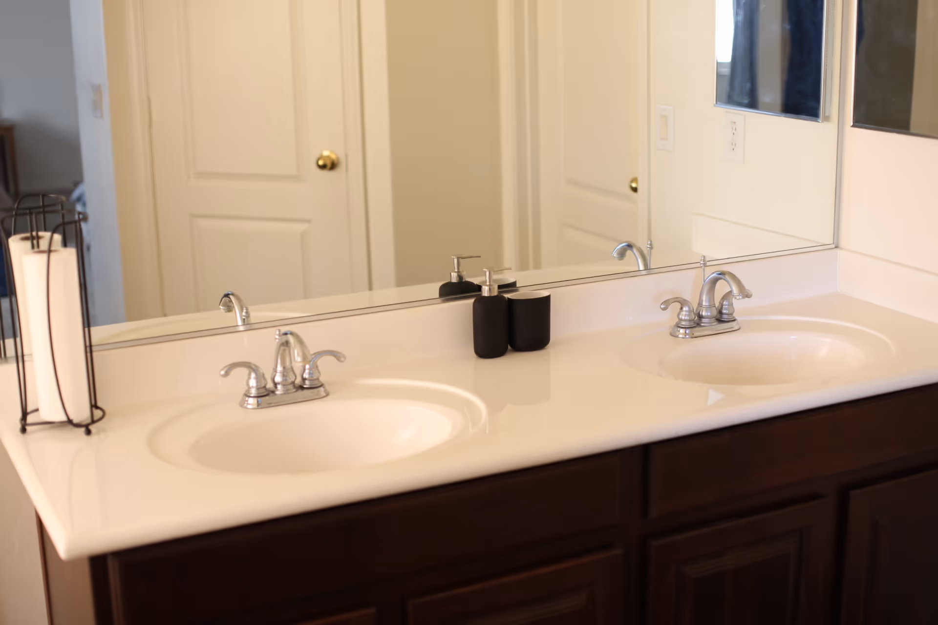 A bathroom countertop with two sinks and chrome faucets. On the left side of the counter, there is a black metal holder with two white rolled towels. In the center, there are two black containers, one with a pump dispenser. A large mirror is mounted on the wall behind the sinks, reflecting a closed white door with a brass doorknob.
