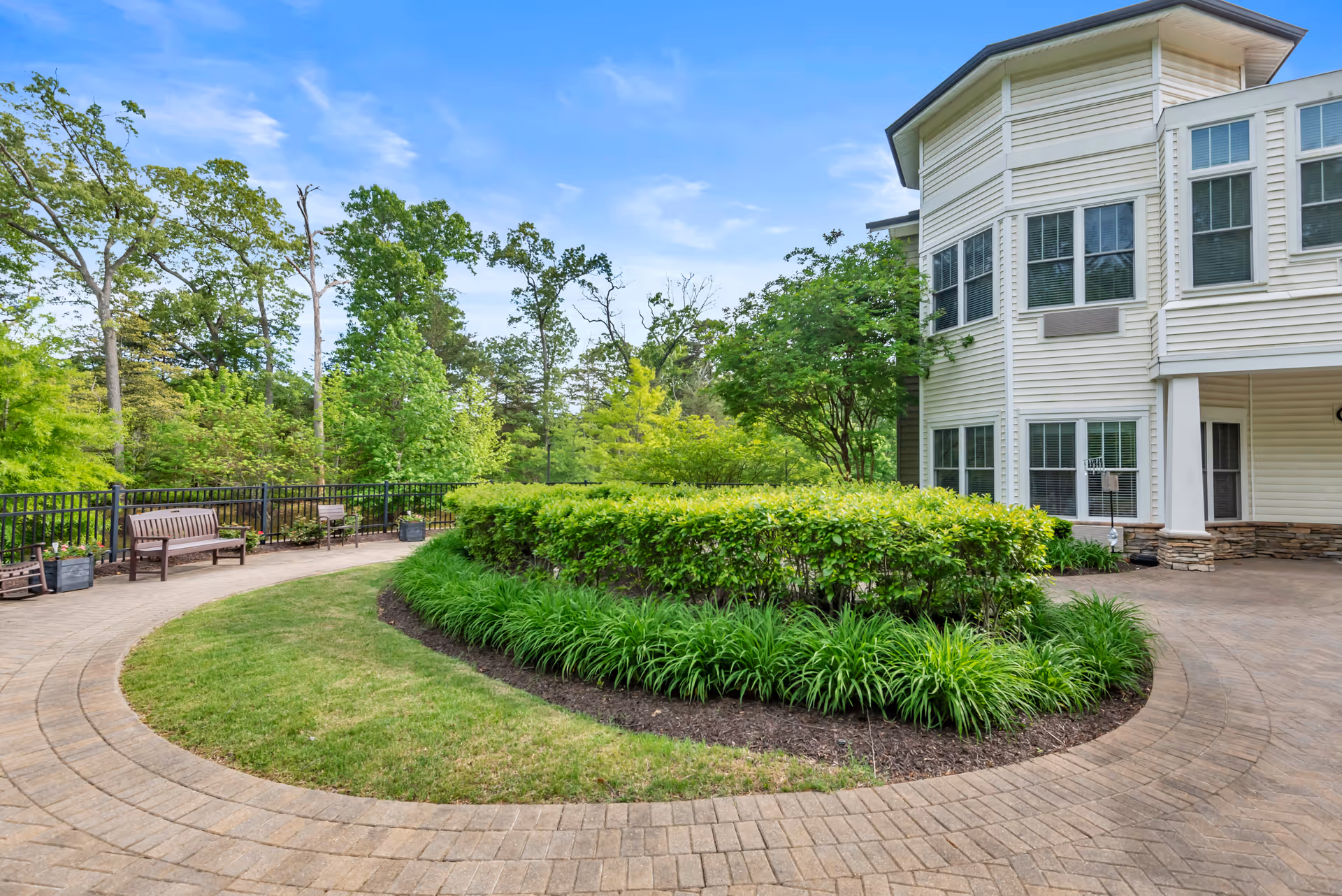 Curved brick pathway surrounding a landscaped garden with green shrubs and grass in front of a white multi-story building with many windows, under a blue sky with some clouds. There are benches along the pathway and trees in the background.