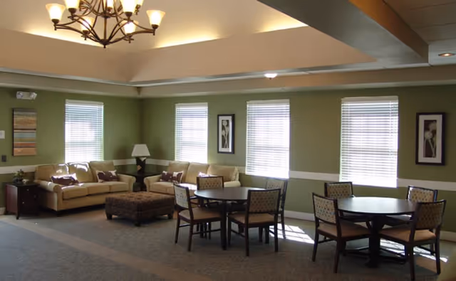 A well-lit living room area with green walls and white trim, featuring two beige sofas with decorative pillows, a large ottoman, two round tables each surrounded by four chairs, three windows with blinds, framed floral artwork on the walls, a side table with a lamp, and a chandelier hanging from a tray ceiling.
