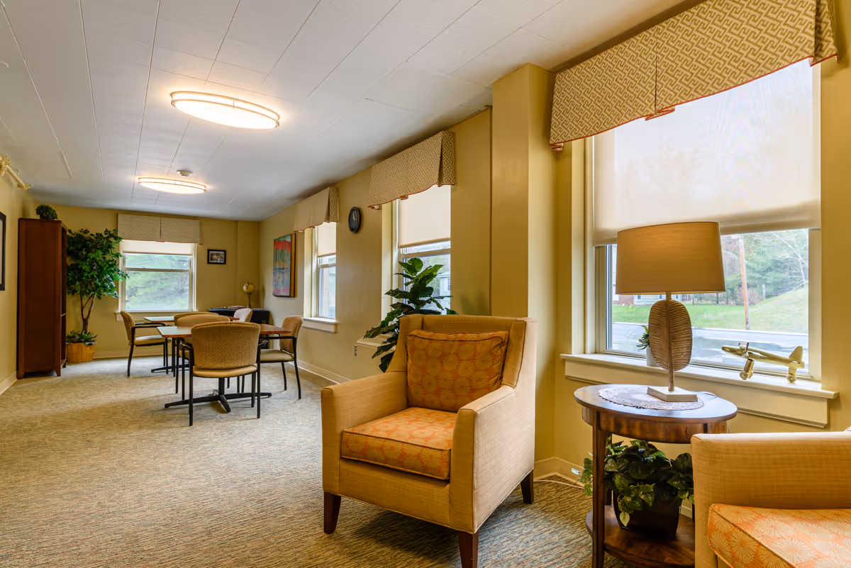 A cozy common area in St. Vincent de Paul Rehabilitation and Nursing Center featuring beige armchairs with patterned cushions, a small round wooden table with a lamp and decorative airplane, several windows with patterned valances, and a table with chairs in the background. The room is warmly lit with ceiling lights and decorated with plants and framed artwork.