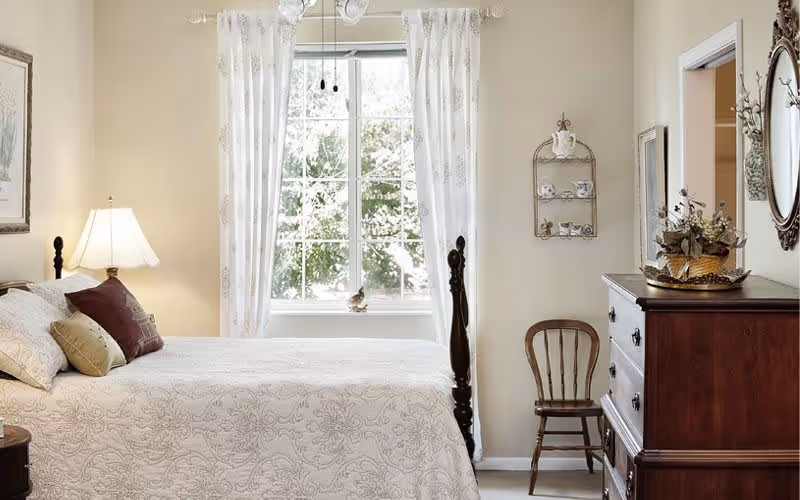 A cozy bedroom with a neatly made bed featuring a white patterned bedspread and multiple pillows. A bedside table with a lamp is on the left side of the bed. A window with white curtains lets in natural light, and a wooden chair is placed against the wall near a wooden dresser with a decorative basket and a mirror above it.