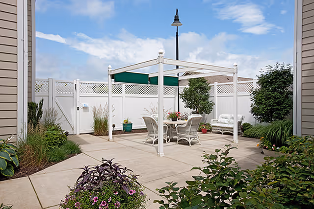 Outdoor patio area with a white pergola structure, a round table with four chairs, and a cushioned bench. The patio is surrounded by a white fence and landscaped with various green plants and flowers. A streetlamp is visible in the background under a partly cloudy sky.