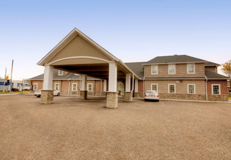 Exterior view of Meadows Assisted Living and Care Campus showing a large covered entrance with stone pillars, a two-story building with multiple windows, and a parking area with two cars.