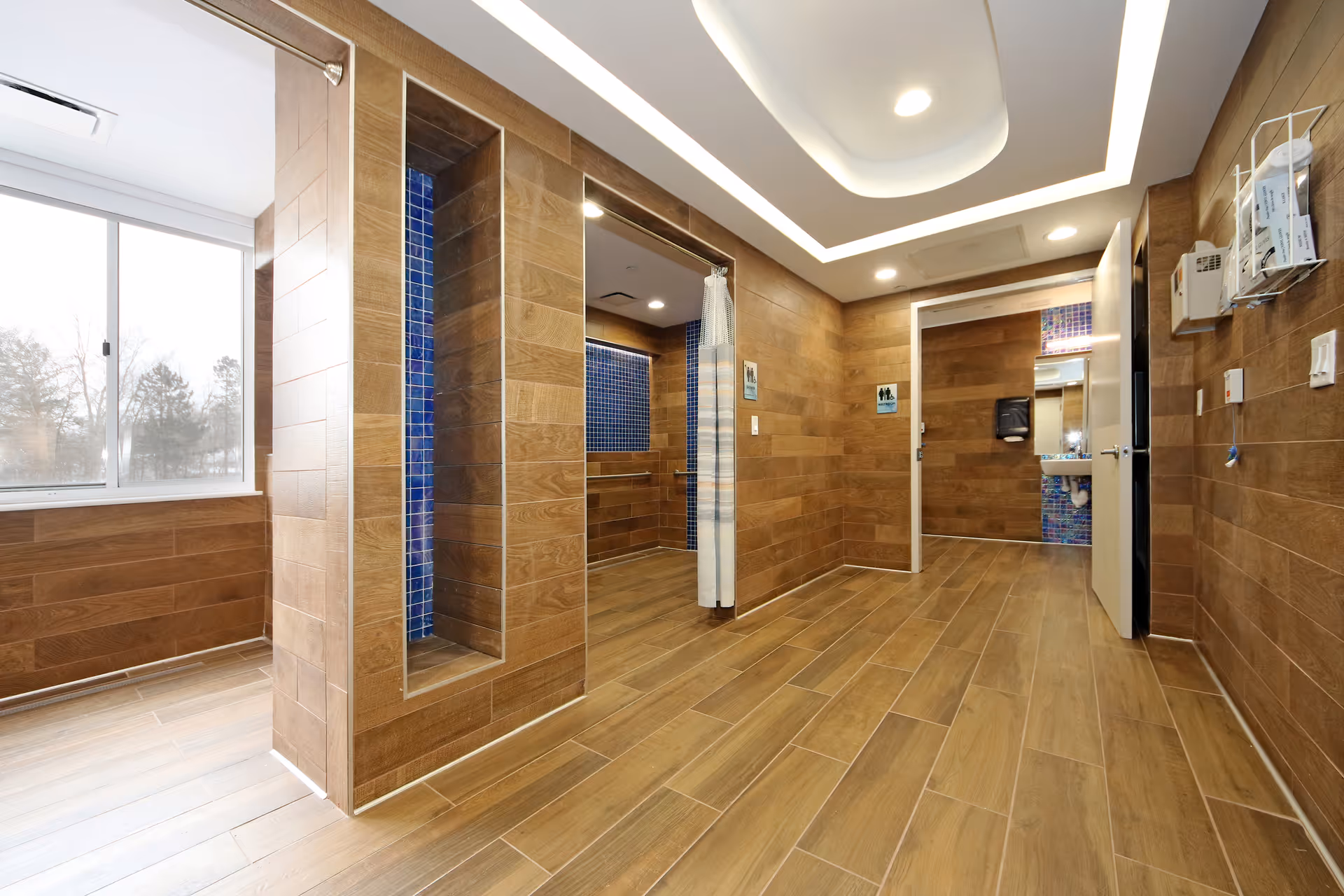A modern wood-tiled restroom and changing area with open shower stalls, a sink room, and a large window.