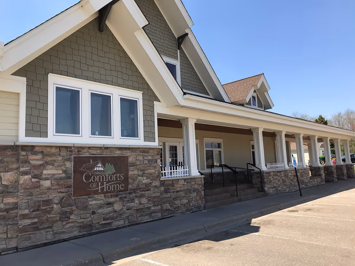 Front exterior of Comforts of Home memory care building showing a stone facade, covered porch with columns, and the facility sign.