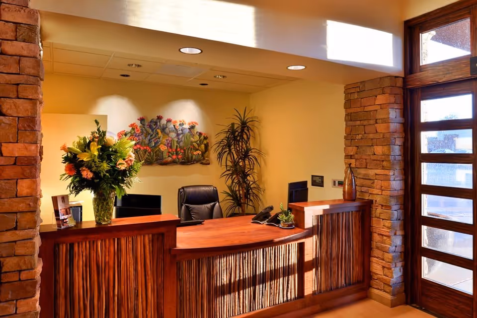 Reception desk area with a wooden counter, vase of flowers, leather chair, potted plant, and stone-accented walls by a glass door.
