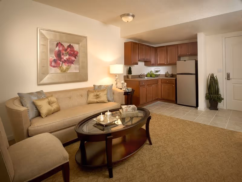 A cozy living area with a beige sofa adorned with decorative pillows, a matching cushioned chair, and a dark wooden oval coffee table with a glass top. Behind the sofa is a framed painting of red flowers. To the right, there is a small kitchen area with wooden cabinets, a countertop, a stainless steel refrigerator, and a tiled floor. A potted plant is placed near the entrance door.