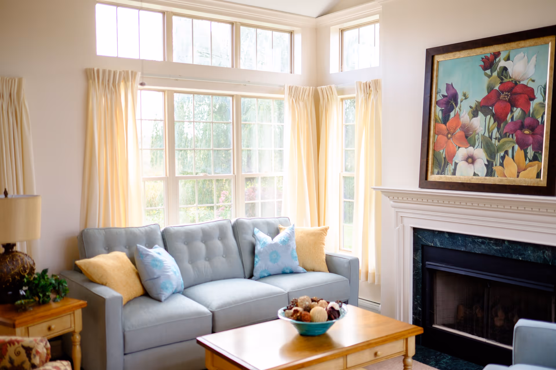 A bright living room with large windows covered by cream-colored curtains, a light blue sofa with yellow and blue patterned pillows, a wooden coffee table with a decorative bowl, a side table with a lamp and plant, and a fireplace with a colorful floral painting above it.
