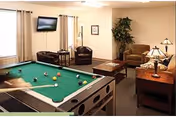 A cozy common area featuring a pool table in the foreground with billiard balls and a cue stick. In the background, there are two leather chairs, a sofa, a coffee table, a side table with a lamp, a potted plant, and a wall-mounted flat-screen TV. The room has light-colored walls and carpeted flooring.