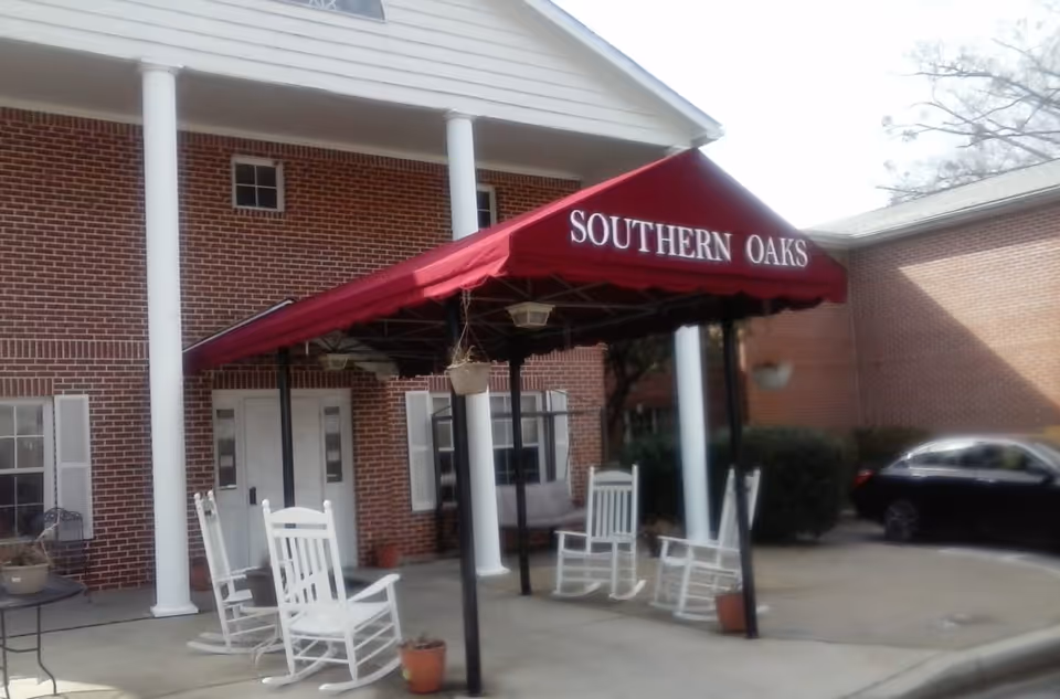 Entrance of a brick building with white columns and a red canopy that reads 'SOUTHERN OAKS'. There are white rocking chairs and potted plants on the concrete area under the canopy. A black car is parked nearby.