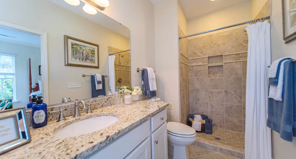 A clean and modern bathroom featuring a granite countertop with a sink, a large mirror above the sink, a toilet, and a tiled walk-in shower with a white curtain. Towels are neatly hung on towel racks and folded inside the shower area. A framed picture is visible on the wall, and part of a bedroom can be seen through the open door.