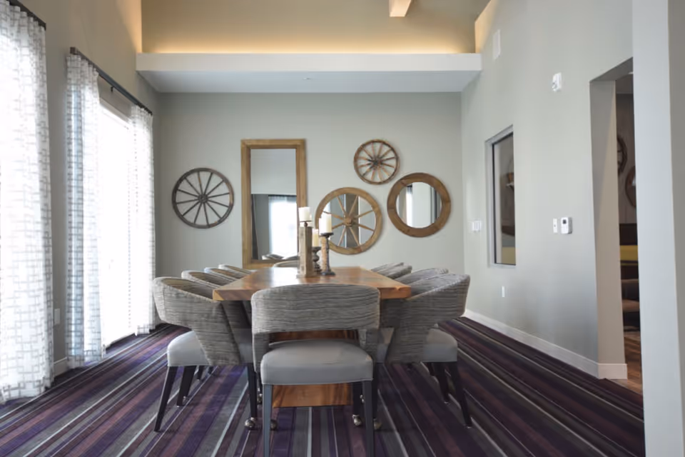 A modern dining room with a wooden table surrounded by eight upholstered chairs. The room features a striped purple carpet and light gray walls decorated with circular wooden wall art and a rectangular mirror. Large windows with sheer patterned curtains allow natural light to fill the space.