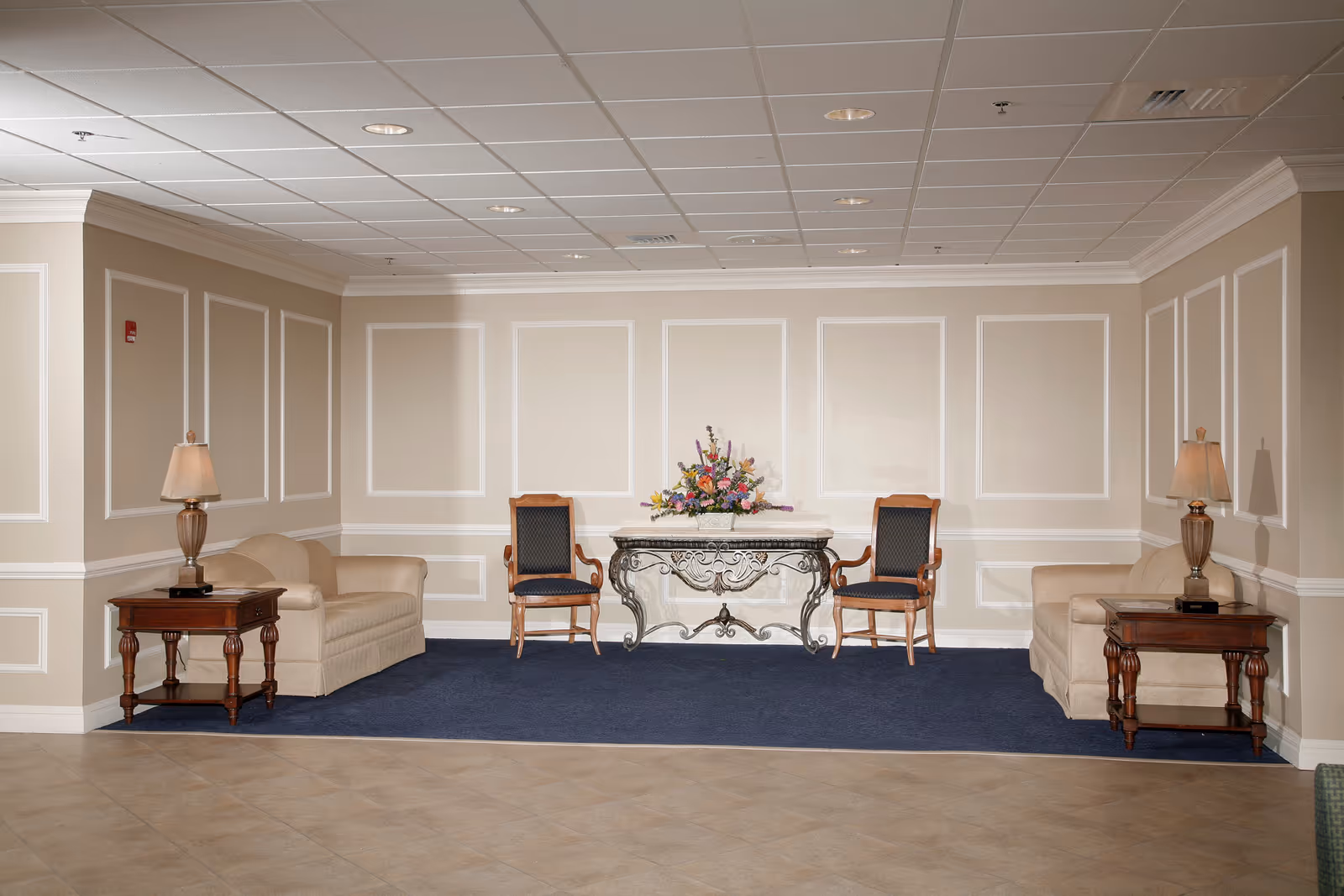A formal interior seating area with two sofas, two chairs, side tables with lamps, and a decorative console table topped with a flower arrangement against paneled walls.