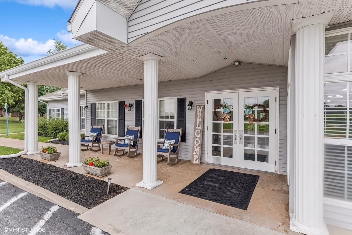 Entrance of Digby Place facility showing a covered porch with white columns, three blue cushioned rocking chairs with small tables, a welcome sign leaning against the wall, double glass doors decorated with wreaths, and flower planters along the edge of the porch.