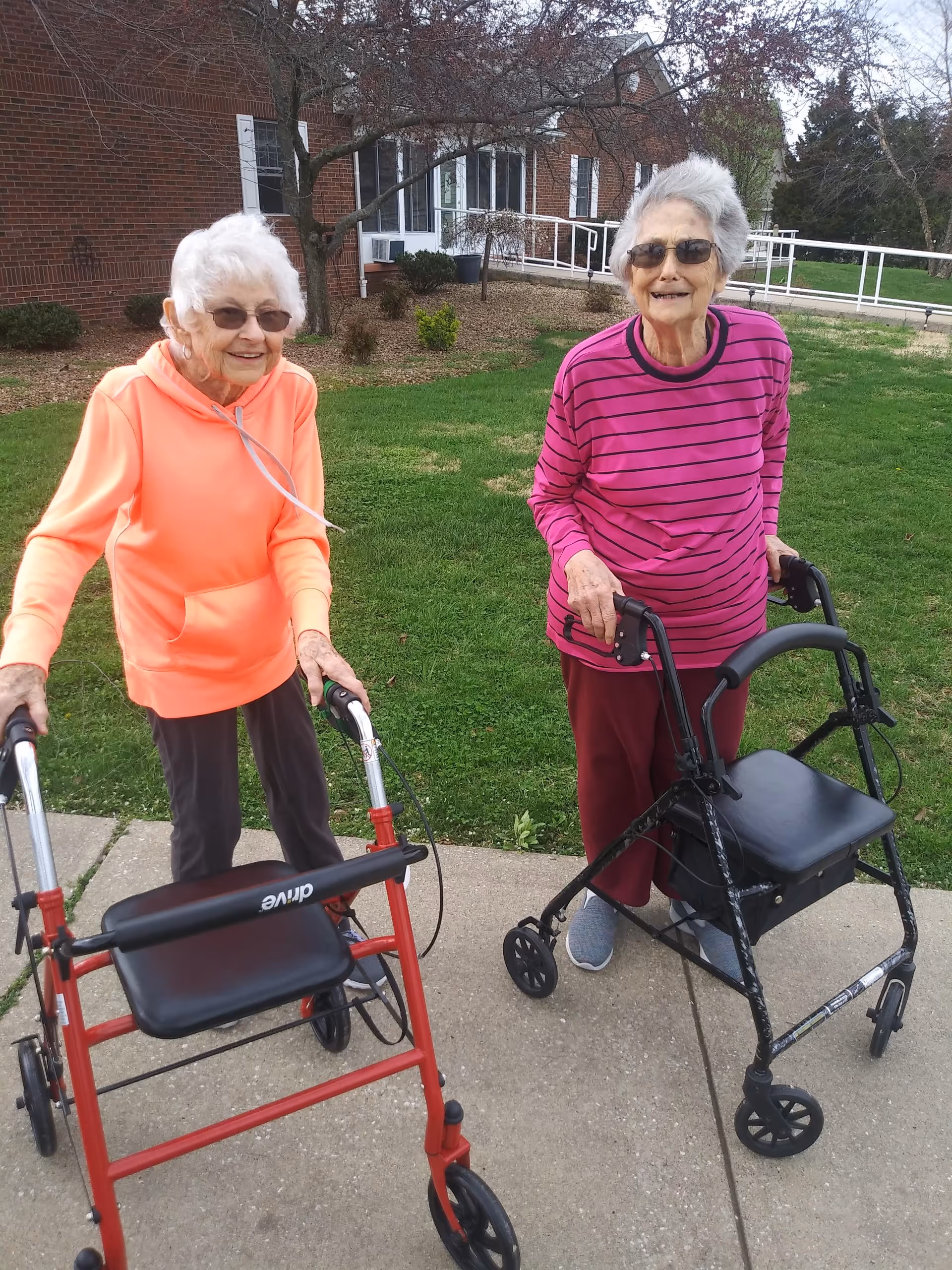 Two elderly women standing outside on a sidewalk in front of a brick building with green grass and trees. Both women are using walkers and wearing glasses. One woman is wearing an orange hoodie and dark pants, while the other is wearing a pink and purple striped long-sleeve shirt and maroon pants. They are smiling and appear to be enjoying the outdoor setting.