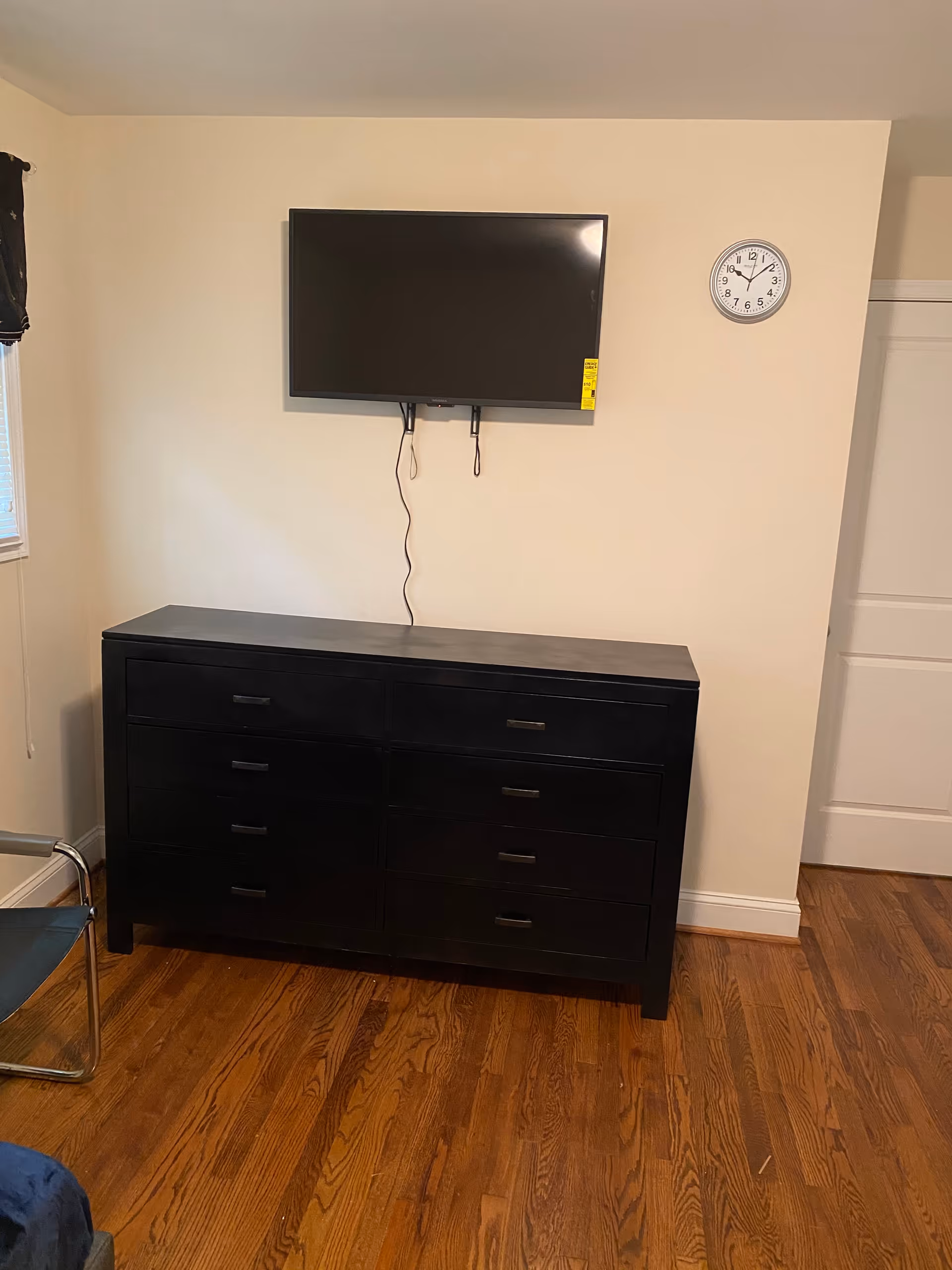 Black dresser against a beige wall with a wall-mounted flat-screen TV and a clock above on a hardwood floor.