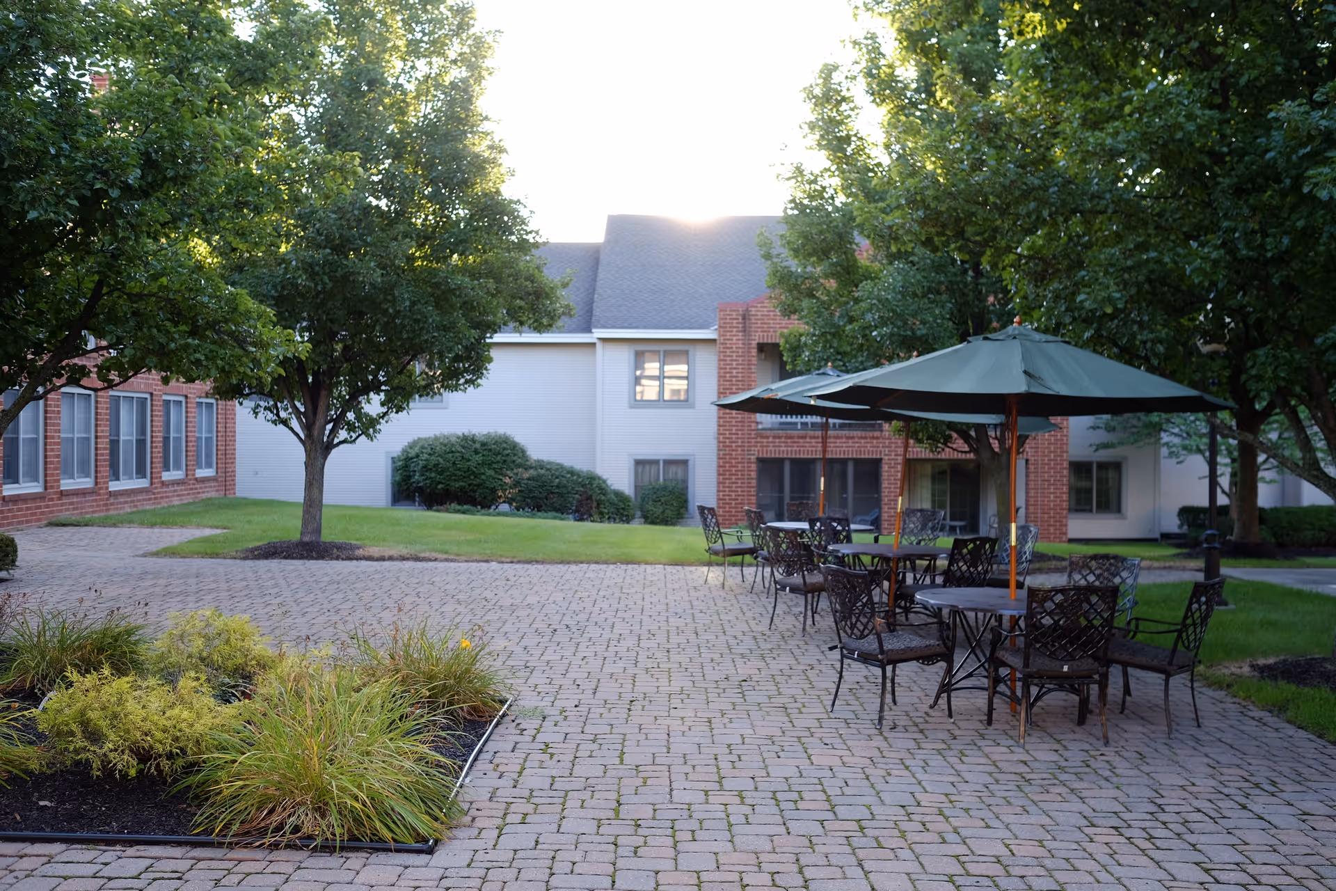 Outdoor courtyard area with brick-paved ground, several metal tables and chairs with green umbrellas, surrounded by green trees and bushes, and a building with red brick and white siding in the background.