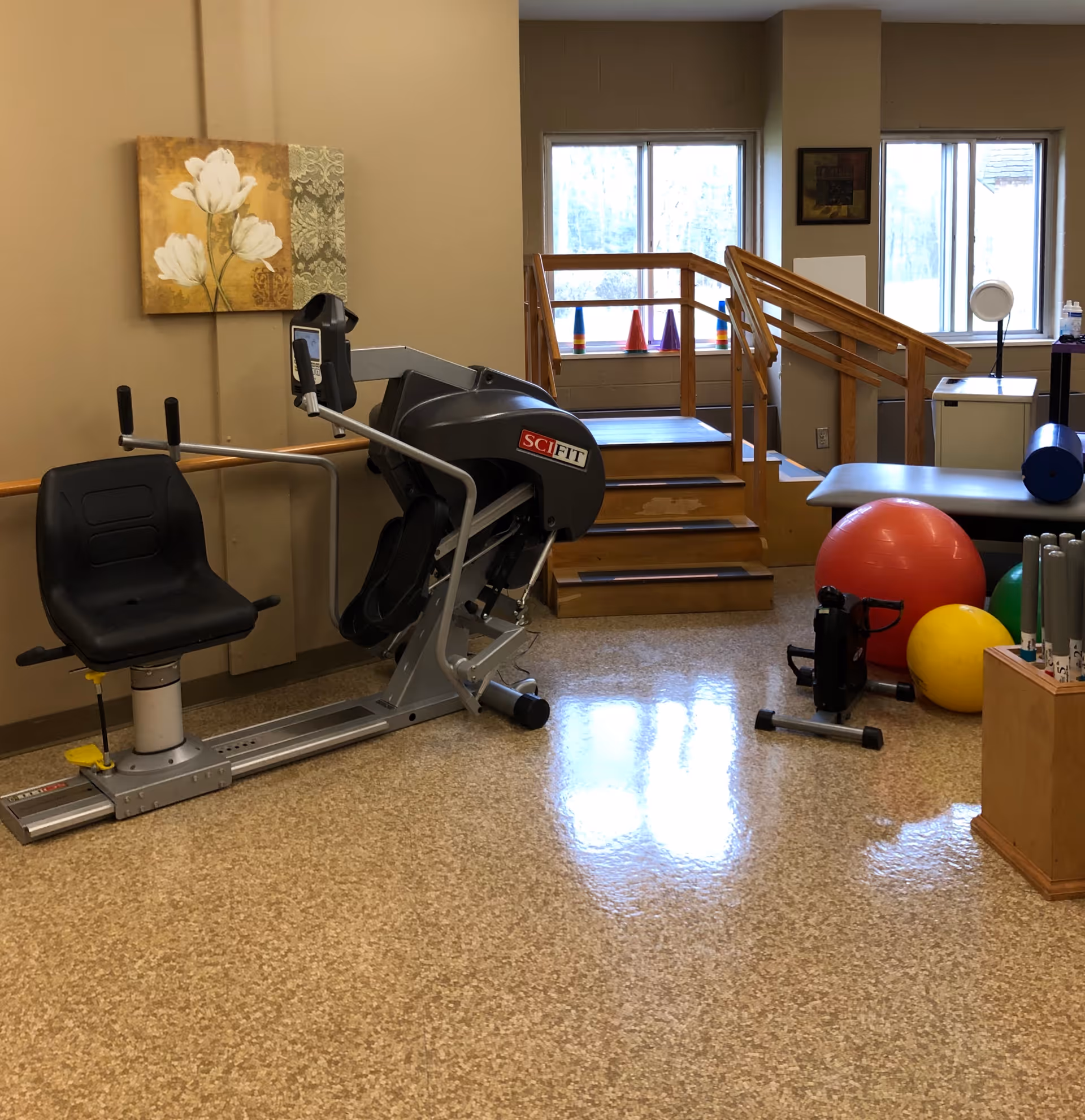 A physical therapy or exercise room with a SciFit exercise machine, a set of wooden stairs with handrails, colorful cones on the windowsill, large exercise balls, a padded bench, and various therapy equipment. The room has beige walls and a polished floor.