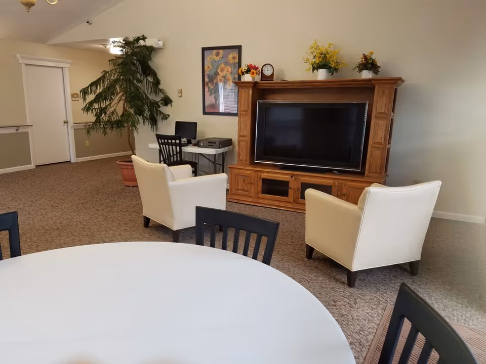 A common area in a senior living facility featuring a round white table with black chairs in the foreground, two beige armchairs facing a large wooden entertainment center with a flat-screen TV. There is a small desk with a computer and printer against the wall, a large potted plant, and a framed sunflower picture on the wall.