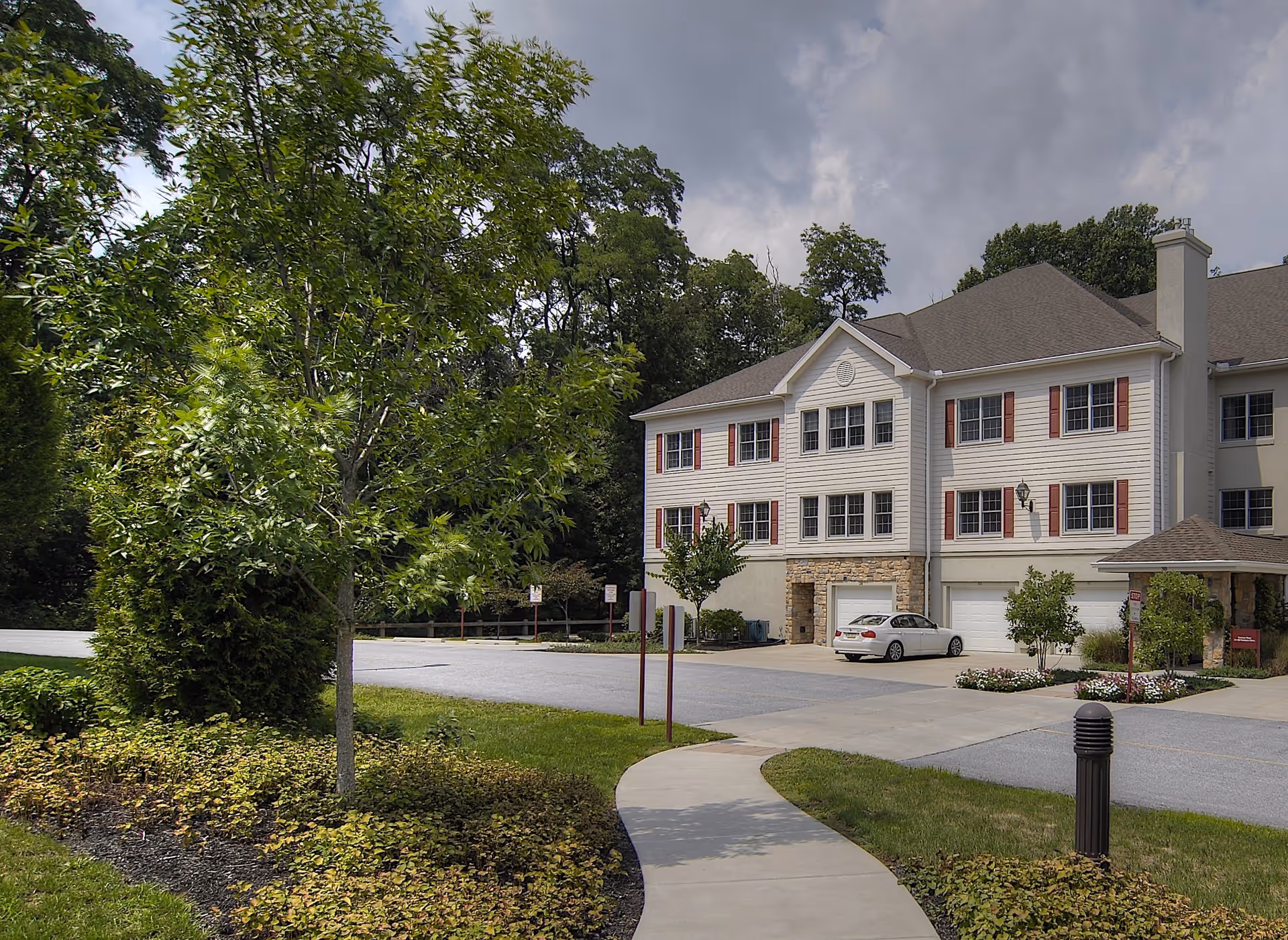 Exterior view of a multi-story residential building with white siding and red shutters, surrounded by trees and greenery. A curved sidewalk leads to the building, and a white car is parked in front of the garages. The sky is partly cloudy.