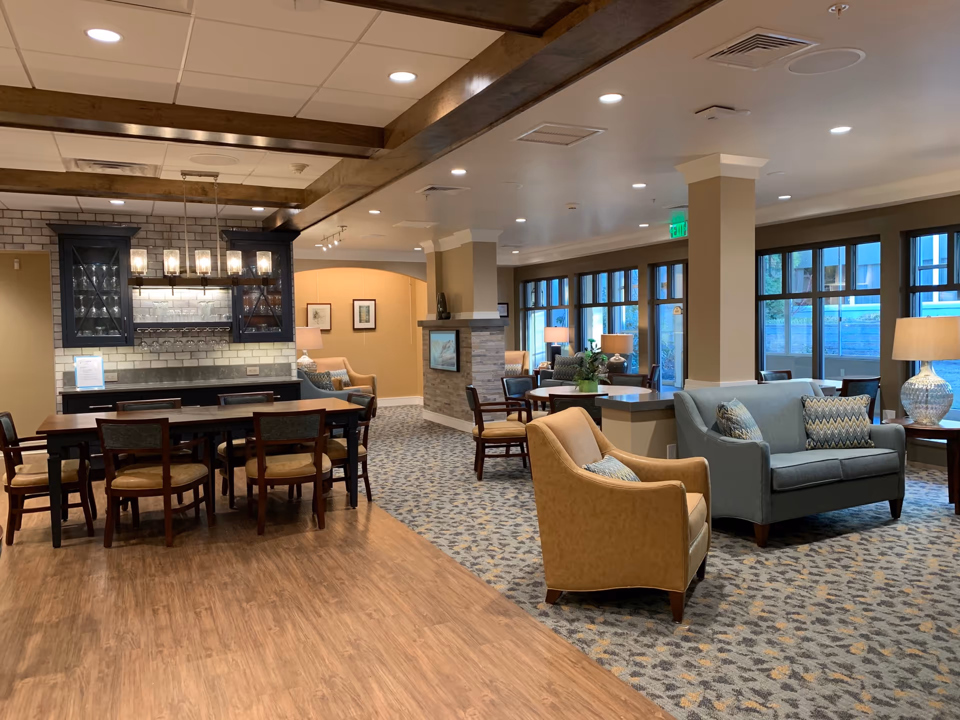 A spacious and well-lit common area in a senior living facility featuring a dining table with chairs on a wooden floor, a sitting area with armchairs and a sofa on carpeted flooring, large windows letting in natural light, and a built-in cabinet with glassware and hanging lights above the dining table.