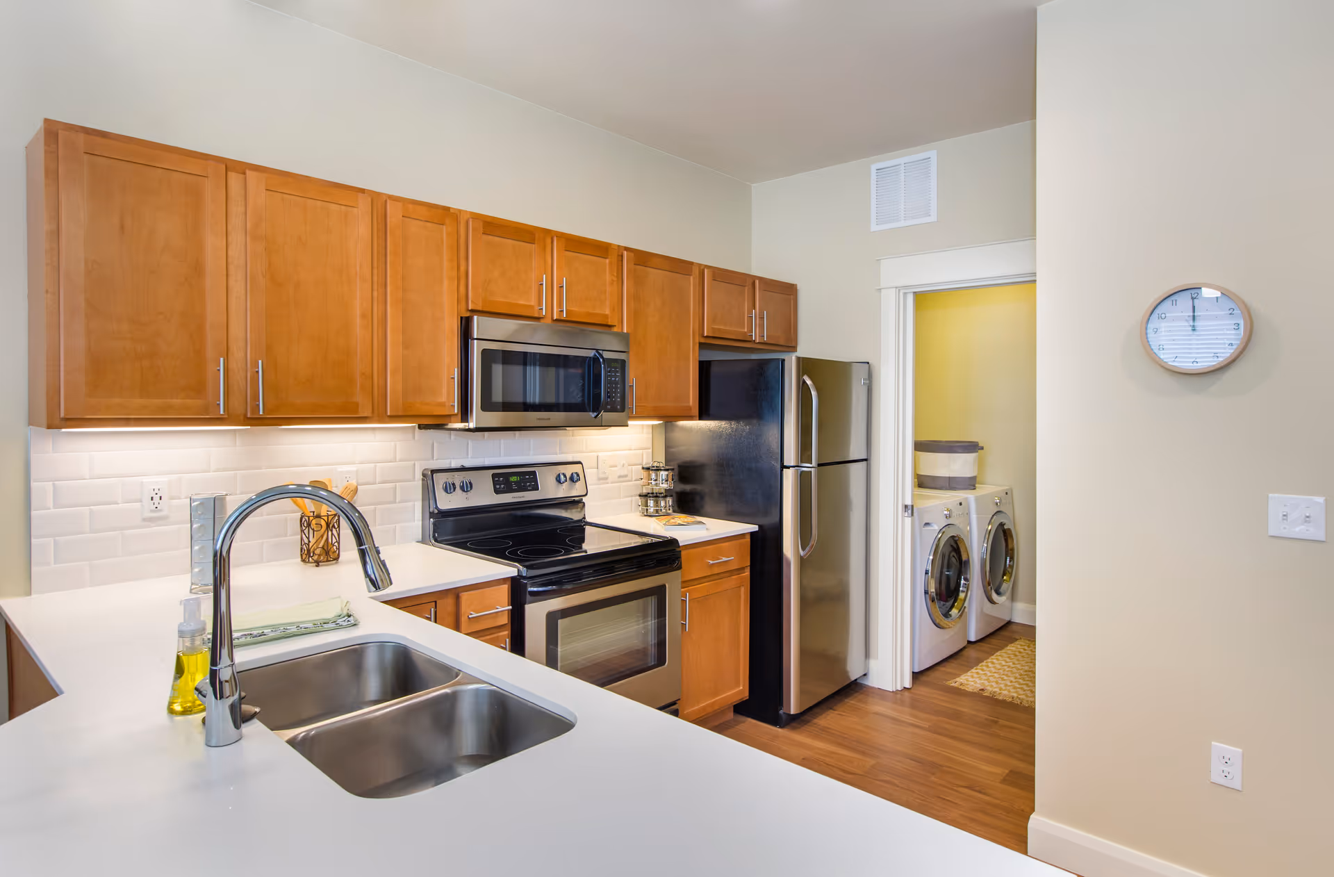 Modern kitchen with white countertops, stainless steel appliances, wooden cabinets and a view into a laundry room.