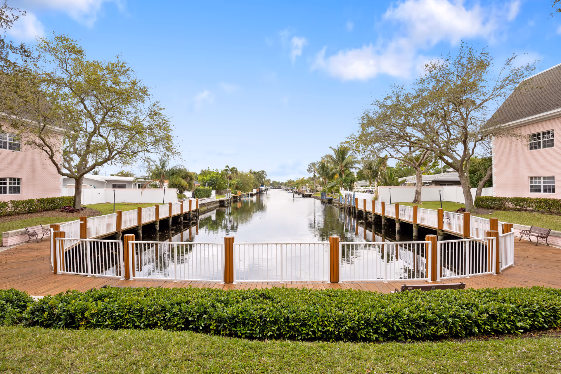 Wooden waterfront boardwalk and fenced dock between two light-pink buildings overlooking a calm canal lined with trees and boats.