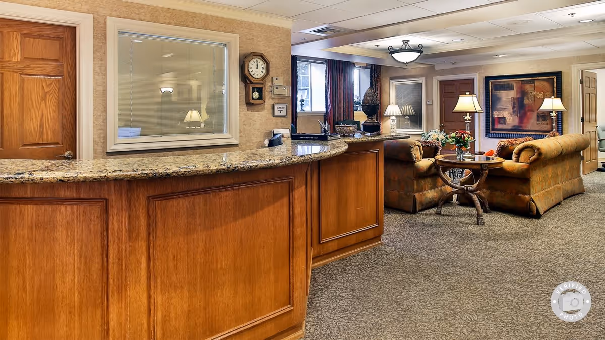 Interior view of a senior living facility reception area with a wooden front desk featuring a granite countertop. Behind the desk is a window with blinds and a wall clock. To the right, there is a cozy seating area with two upholstered sofas, a round wooden coffee table with a flower arrangement, several table lamps, and framed artwork on the wall. The room is carpeted and has soft lighting.