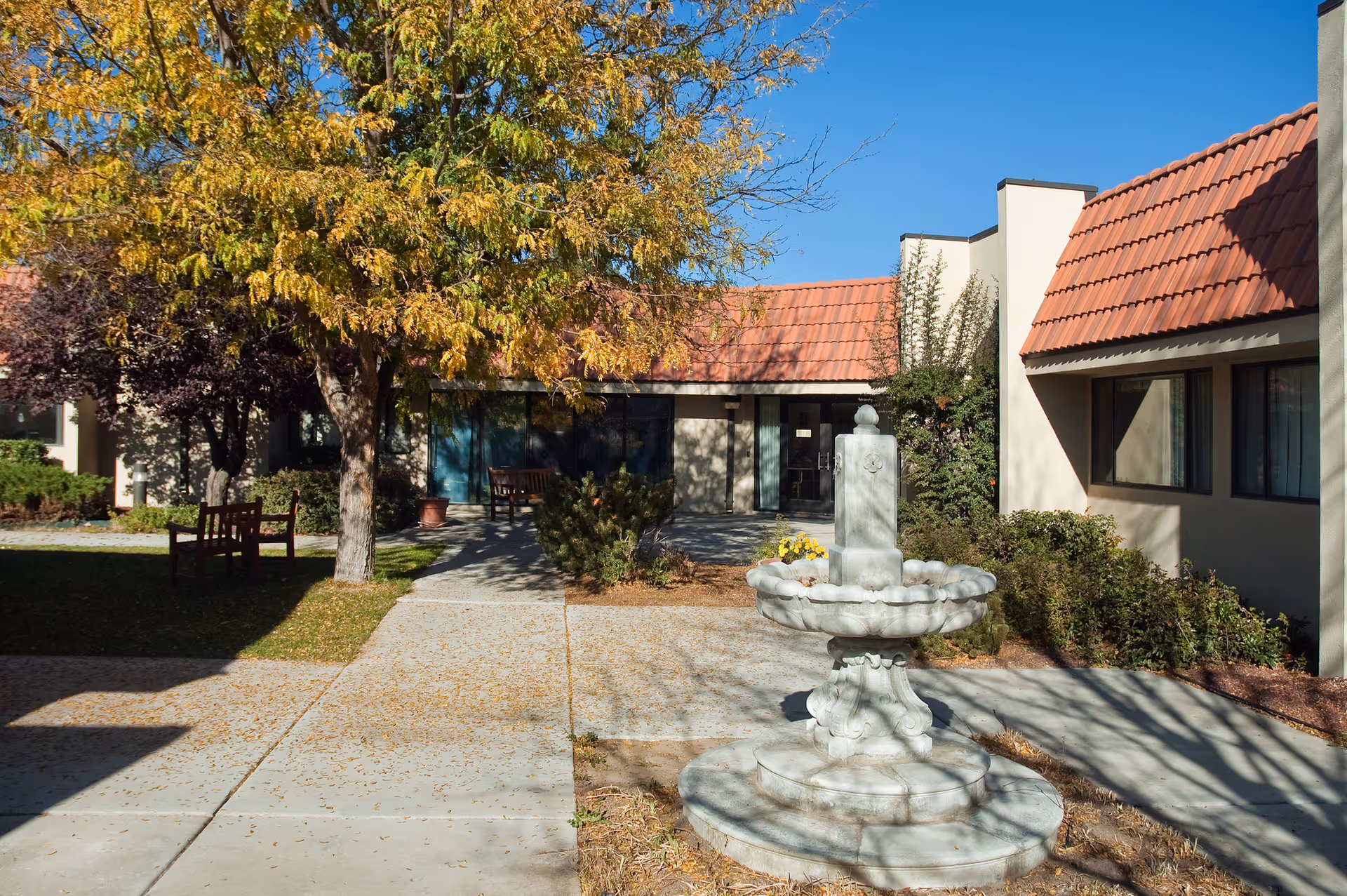 Outdoor courtyard area of a senior living facility with a stone fountain in the foreground, a tree with yellowing leaves, benches, and buildings with red-tiled roofs under a clear blue sky.
