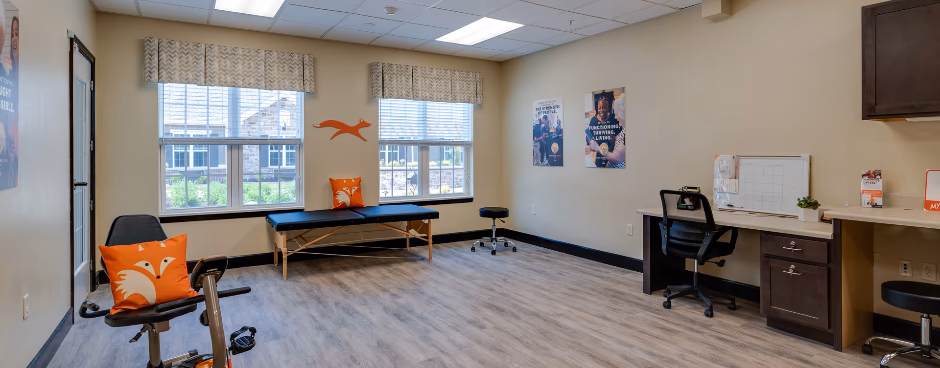 A bright, spacious room with large windows covered by patterned valances, featuring a black exercise bike with an orange fox pillow, a black padded bench with a matching fox pillow, a black rolling stool, and a desk area with a black office chair, whiteboard, and small plant. The walls are beige with motivational posters and a decorative orange fox wall art.