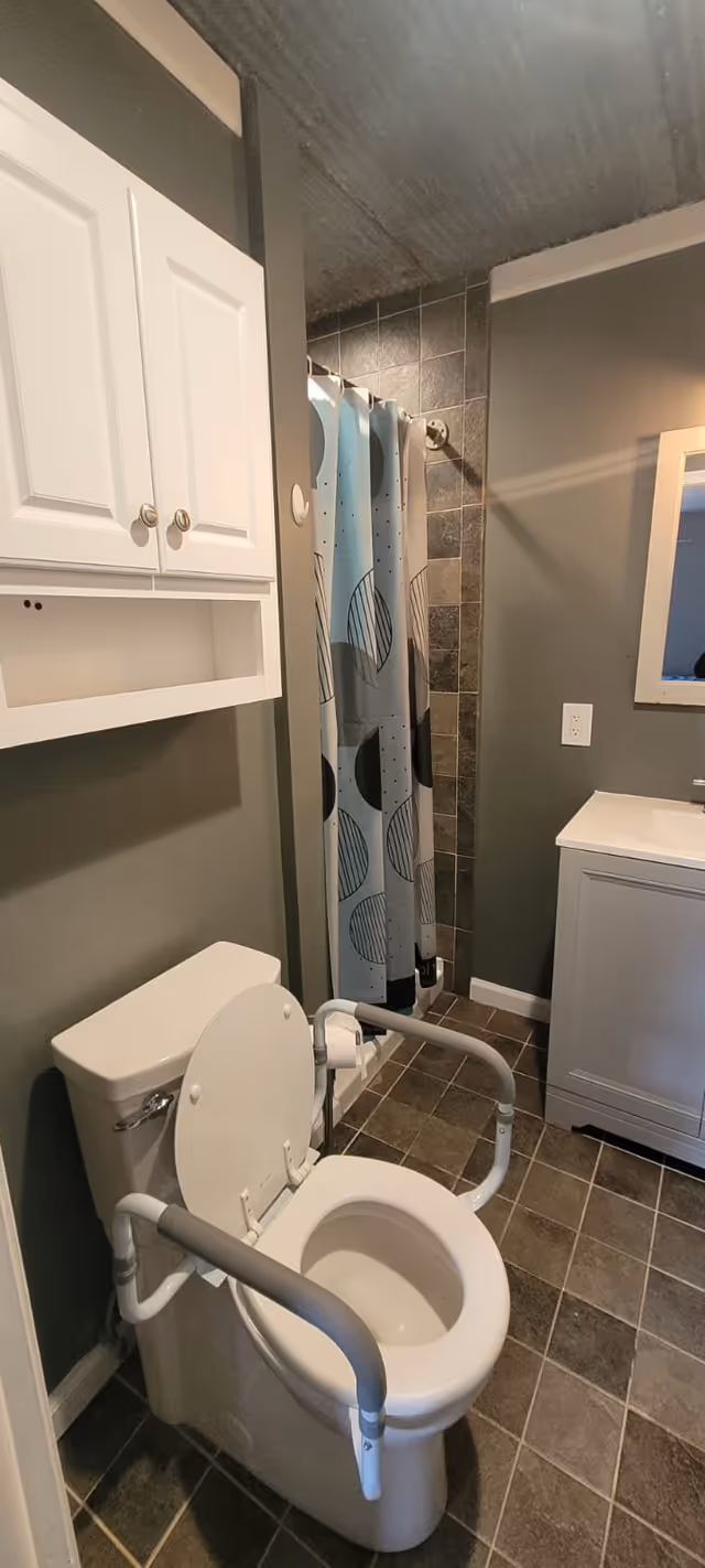 A bathroom with a white toilet equipped with safety rails on both sides, a white cabinet mounted on the wall above the toilet, a shower area with a curtain featuring a geometric pattern, a white sink vanity with a mirror above it, and tiled floor and walls.