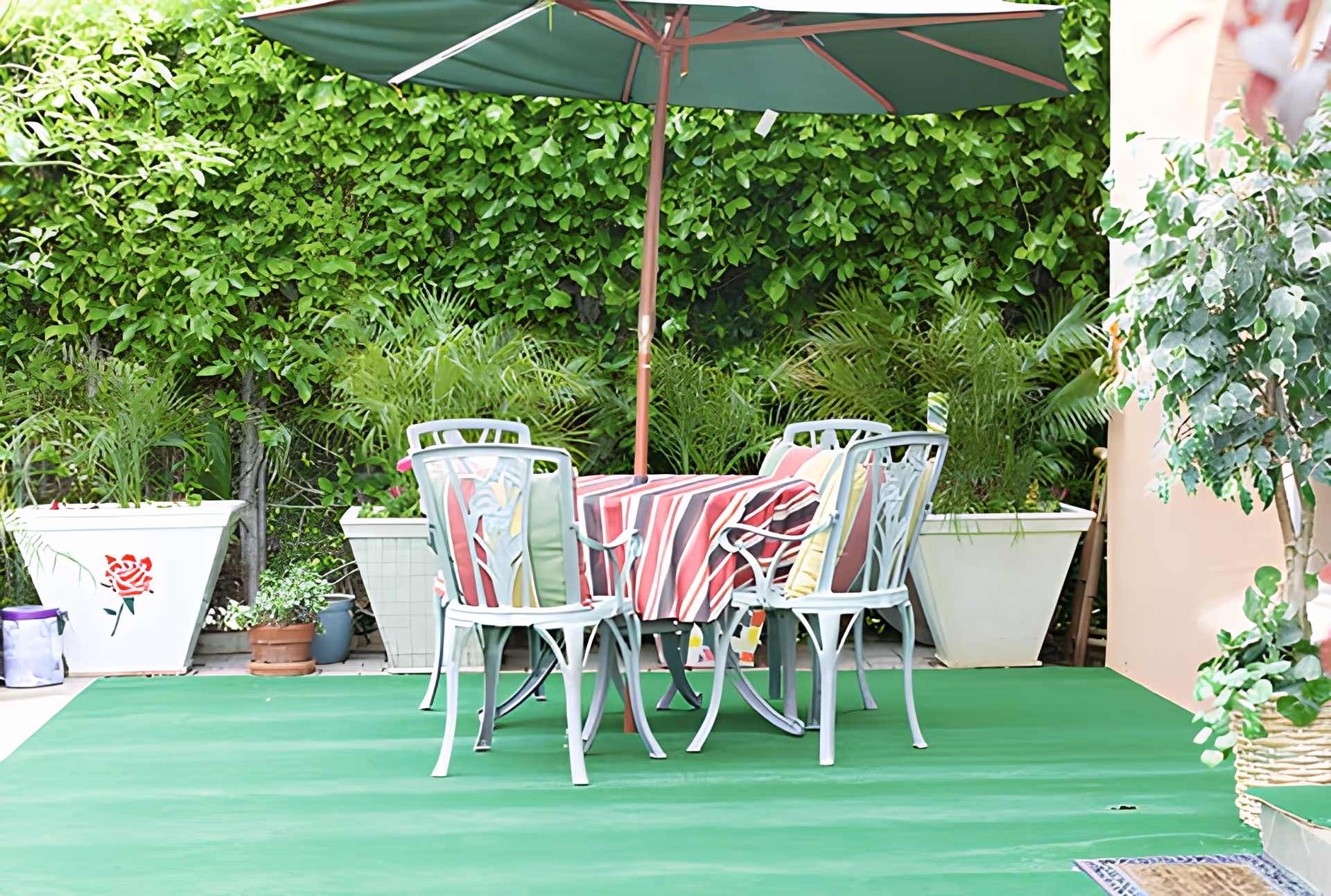 Outdoor patio area with a round table covered by a red and white striped tablecloth, surrounded by four white chairs with colorful cushions. A large green umbrella provides shade over the table. The background features lush green plants and large white planters.