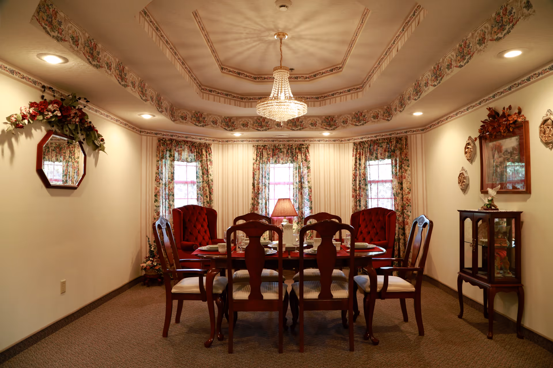 A traditional dining room with a wooden dining table set for six, surrounded by six wooden chairs with cushioned seats. The room features floral curtains on three windows, a chandelier hanging from a decorative ceiling, floral wallpaper border near the ceiling, and a small cabinet with decorative items on the right wall. There is also a wall mirror with floral decoration on the left wall.
