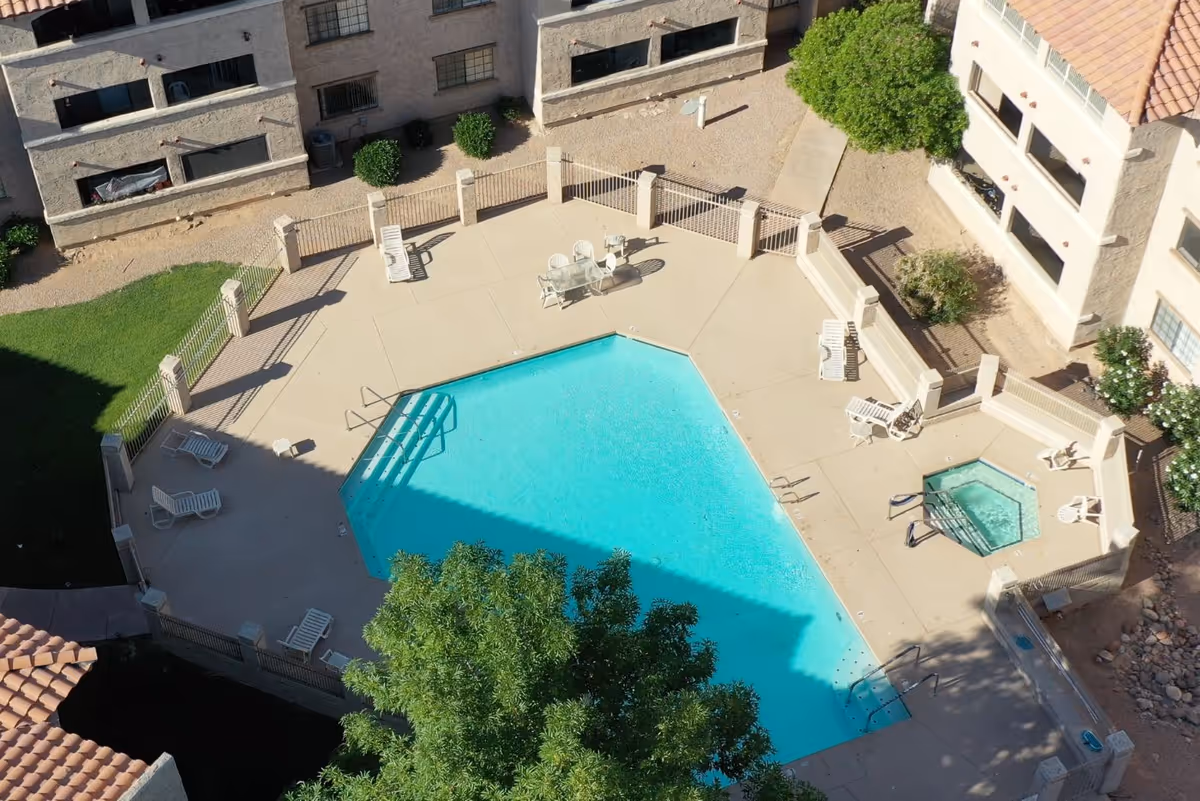 Aerial view of a fenced outdoor swimming pool area with a hot tub, surrounded by lounge chairs and tables with chairs. The pool area is adjacent to a multi-story building with balconies and windows, and there are trees and landscaping around the pool.