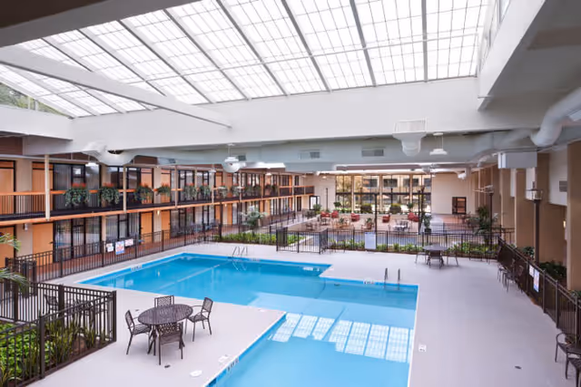 Indoor swimming pool area with clear blue water surrounded by a safety fence. There are several round tables with chairs around the pool. The area is enclosed by two levels of rooms with balconies and potted plants. A large skylight ceiling allows natural light to illuminate the space.