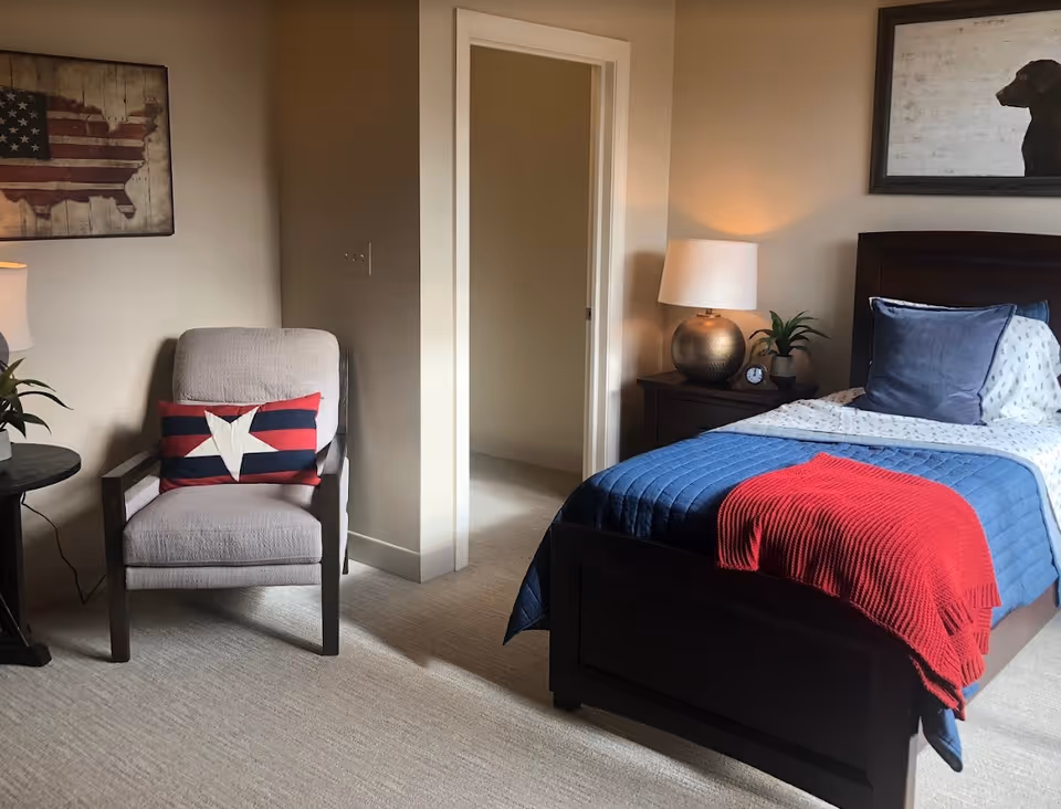 Well-lit bedroom with a single bed dressed in blue and red bedding, a bedside table with lamp, and an armchair with a star pillow.