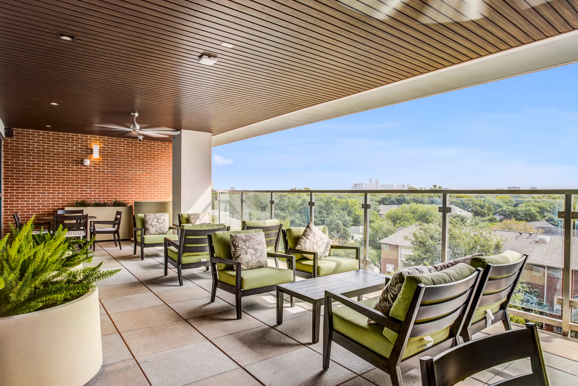 A spacious outdoor balcony area with modern green cushioned chairs and sofas arranged around low tables. The balcony has a glass railing offering a view of trees and distant buildings under a clear blue sky. The ceiling is wooden with recessed lighting and a ceiling fan, and there is a brick wall on one side with additional seating and a potted plant in the foreground.