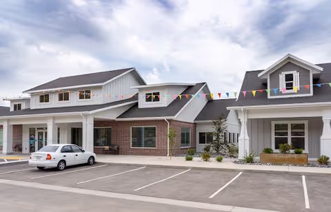 Exterior view of Petersen Farms Assisted Living facility showing a modern building with a combination of brick and light-colored siding, a parked white car in front, and colorful triangular pennant flags strung across the front of the building under a cloudy sky.
