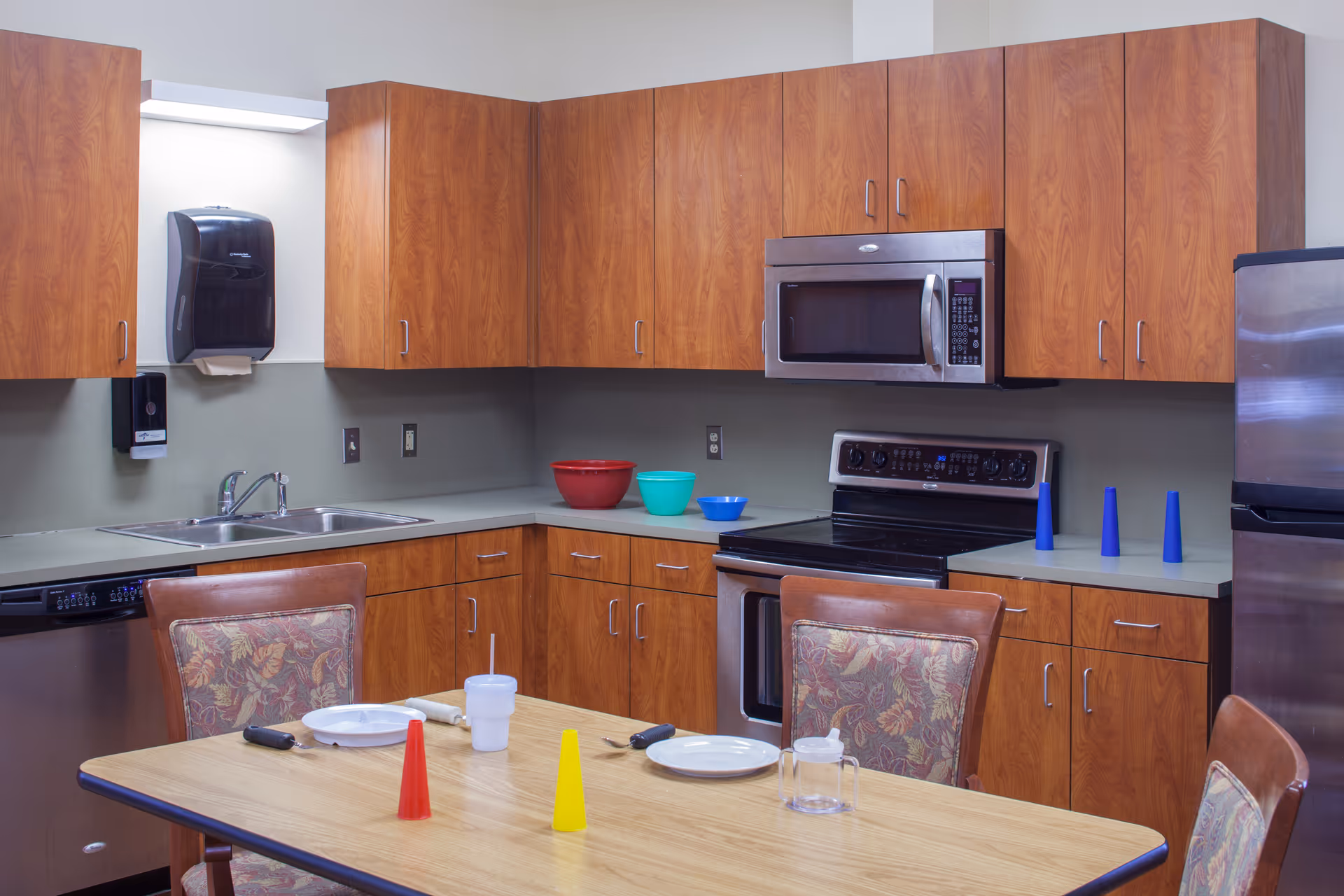 A kitchen area with wooden cabinets, a stainless steel microwave, stove, dishwasher, and refrigerator. A table with four chairs is in the foreground, set with plates, utensils, a cup, and colorful plastic cones. Bowls and additional cones are on the countertops.
