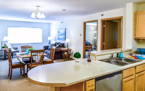 Interior view of a senior living facility showing a kitchen counter with a sink and dishwasher in the foreground, and a dining area with a wooden table and chairs. Beyond the dining area is a living room with a sofa, armchair, side tables, lamps, and a large window letting in natural light. Two doorways lead to other rooms.