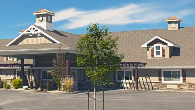 Exterior view of a single-story assisted living facility building with a large covered entrance, clock tower, and multiple windows. There is a small tree in the foreground and a clear blue sky above.