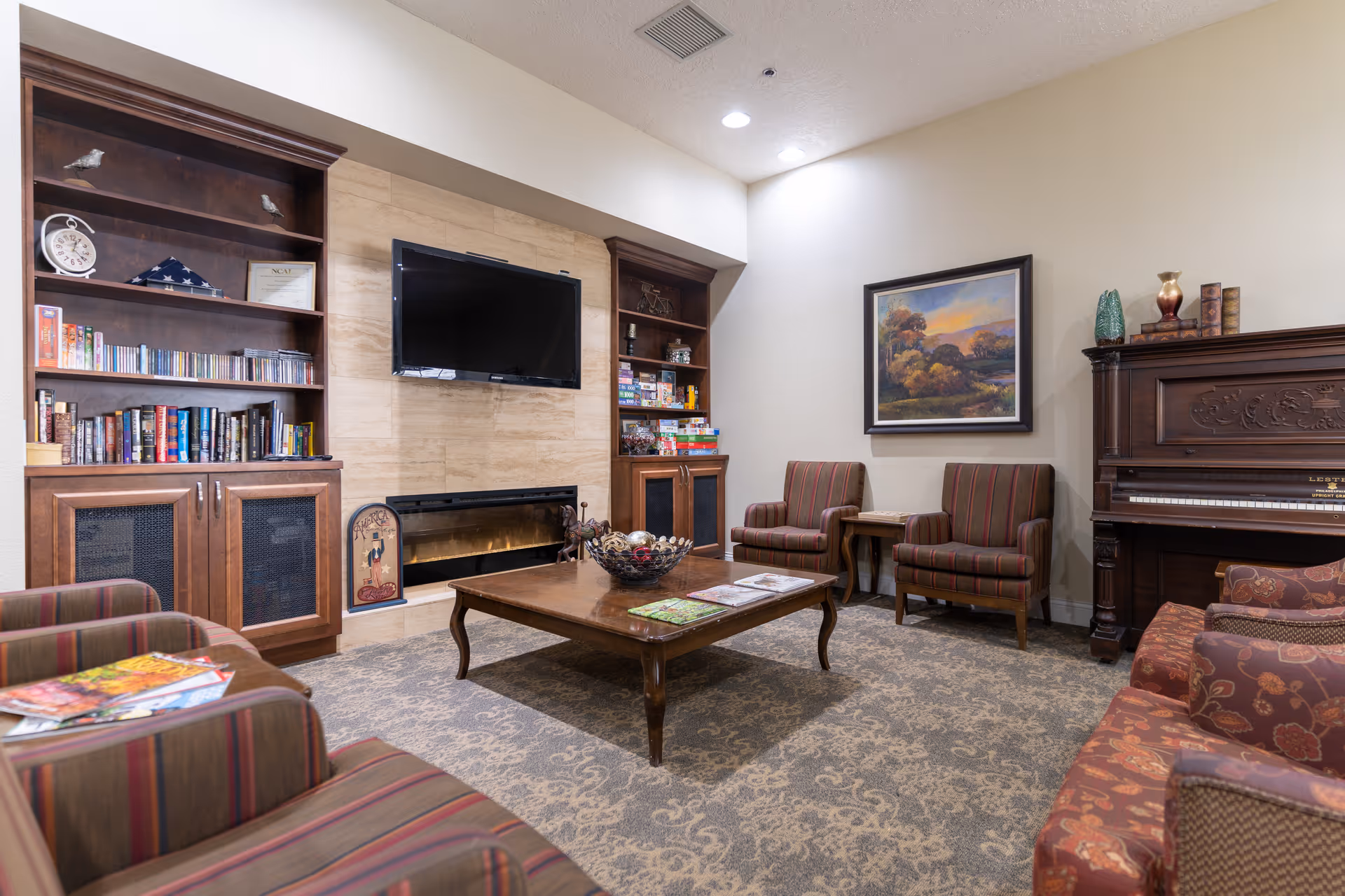 A cozy living room area with a large wooden coffee table in the center, surrounded by striped and floral upholstered armchairs and sofas. The room features built-in wooden shelves filled with books, CDs, and board games, a flat-screen TV mounted above a fireplace, a framed landscape painting on the wall, and an upright piano with decorative items on top.